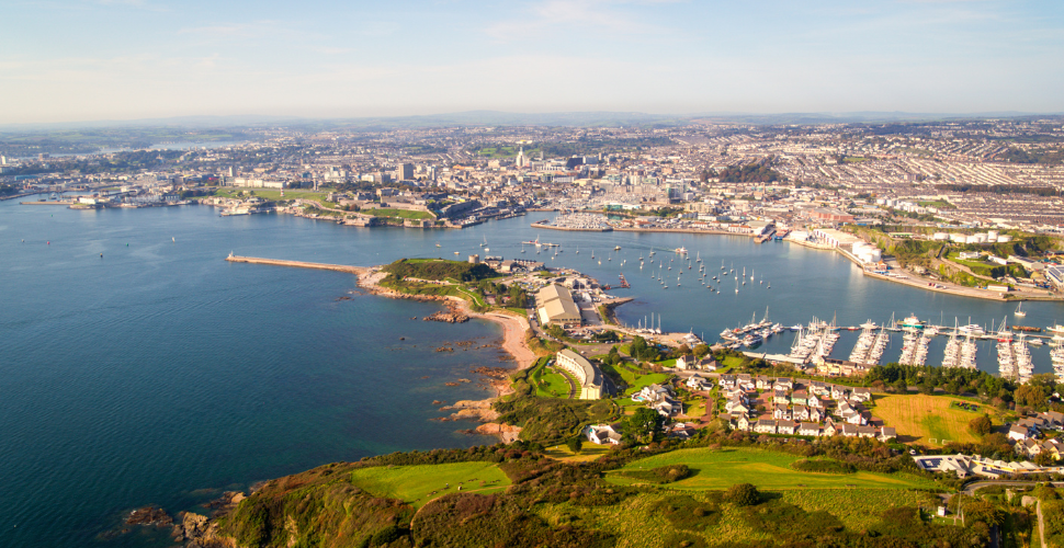 Aerial view of Plymouth’s coastline and harbour with marinas, green headland and the city in the background.